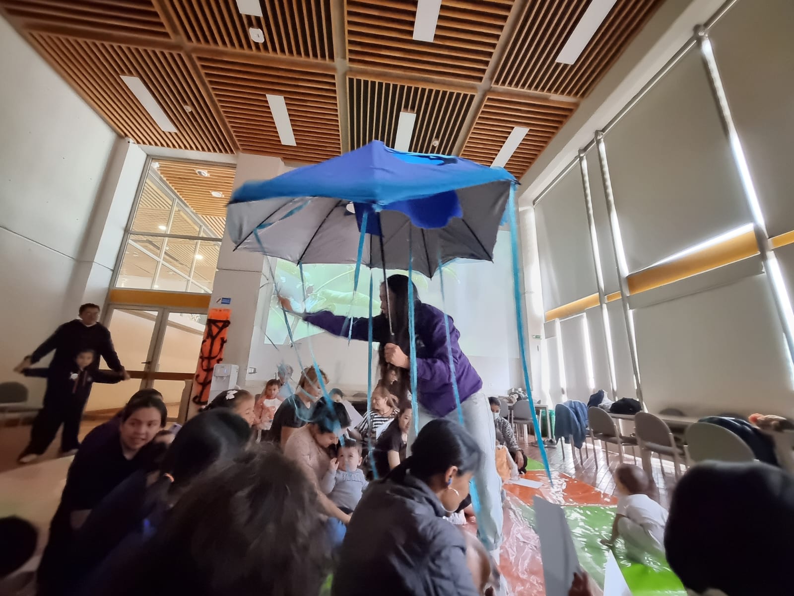 niños y niñas en medio de una obra de teatro mientras interactúan con un paraguas azul.