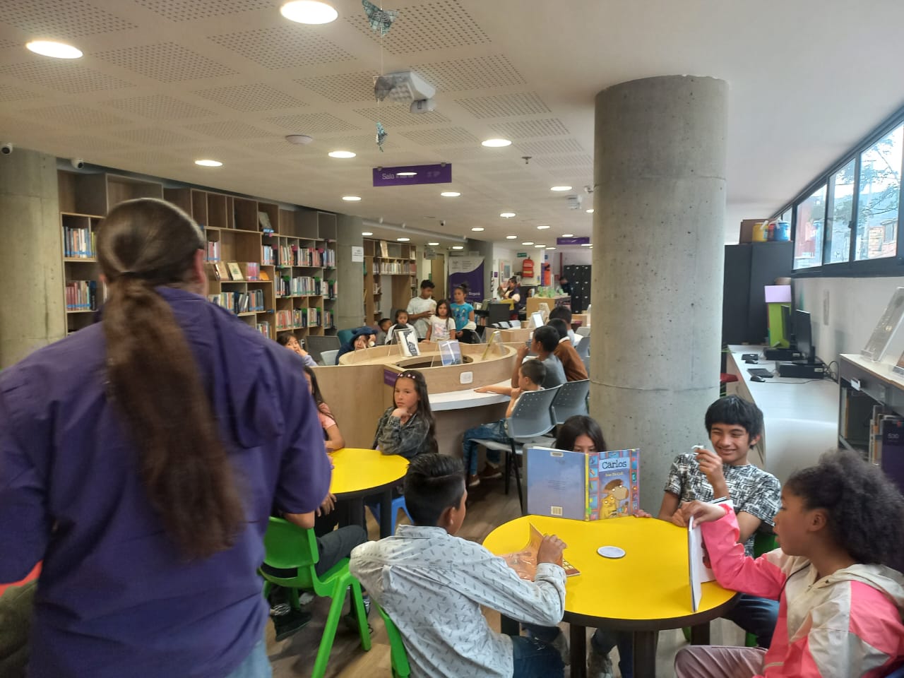 grupo de niños en un salón de biblioteca mientras un docente lee un libro en voz alta.