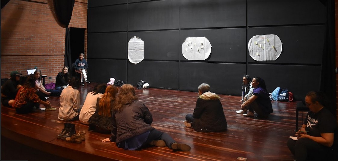 grupo de mujeres sentadas en el suelo de la tarima de un teatro mientras conversan