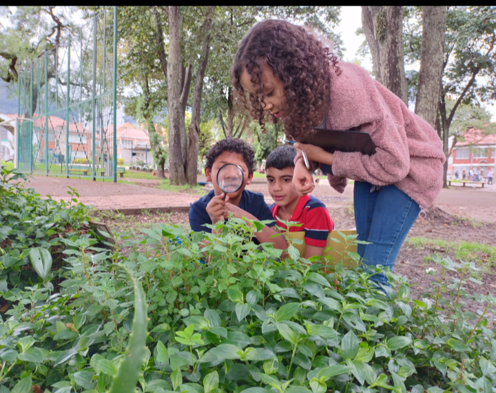 dos niños observando una planta con lupa mientras la maestra los acompaña