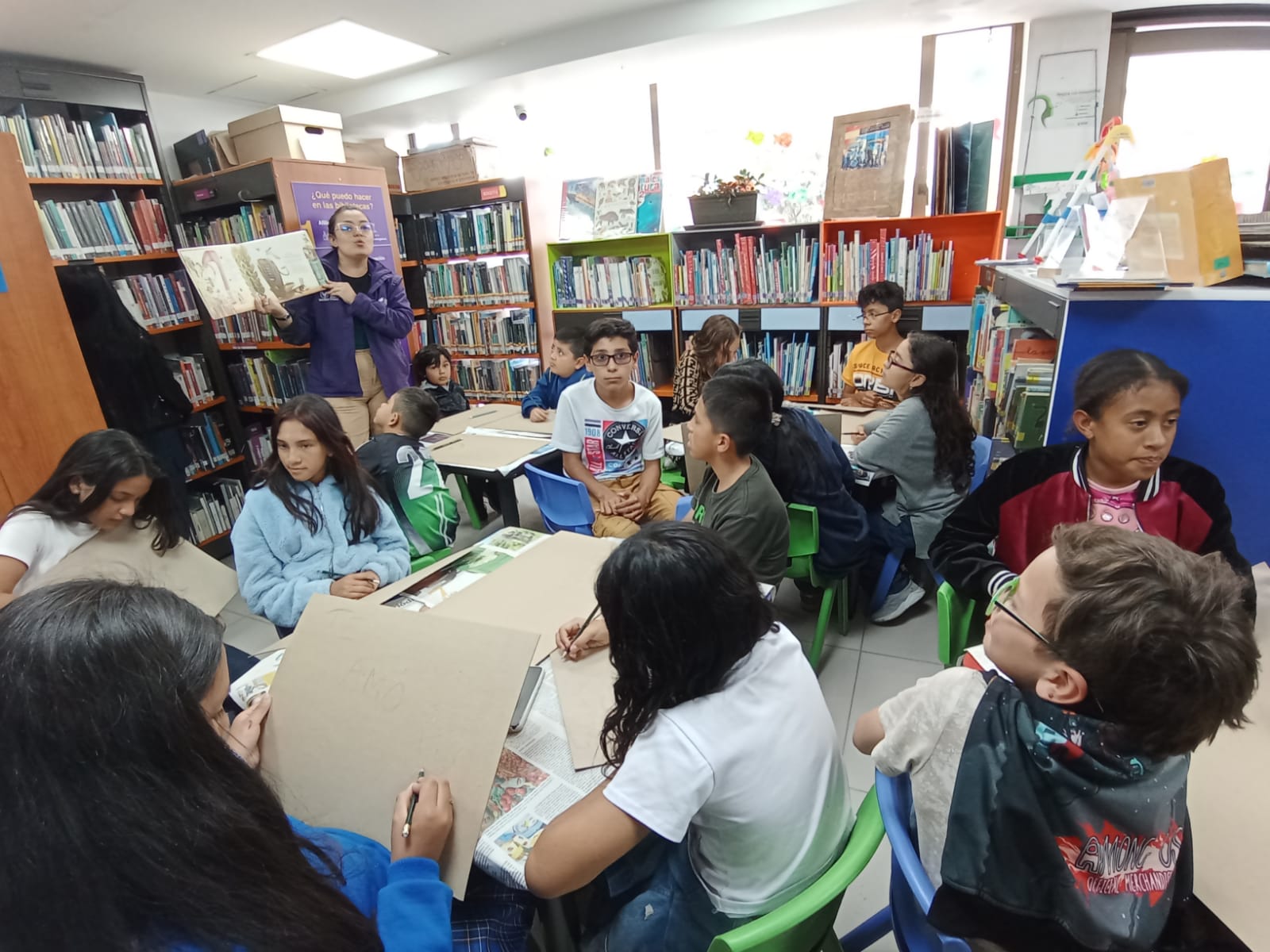 grupo de niños y niñas sentados en varias mesas mientras la docente lee un libro en voz alta 