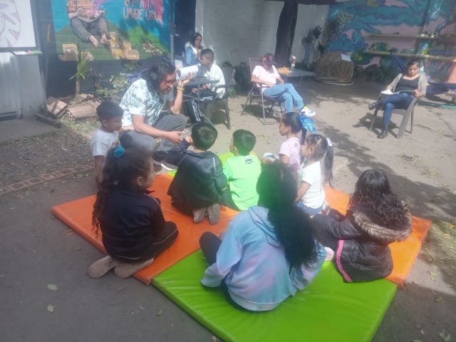 grupo de niños y niñas al aire libre escuchando una lectura grupal por parte de un docente